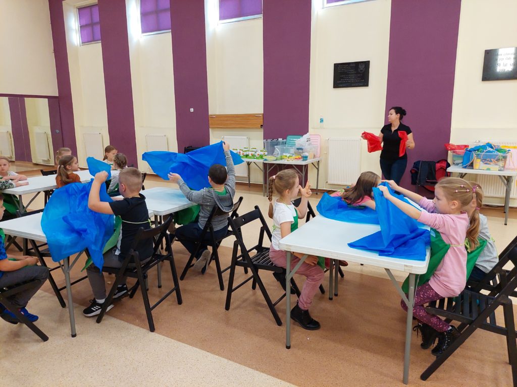 Aula, dzieci siedzące przy 2 białych stołach przykrywają je niebieską folią. W tle Pani Daria trzymająca w rękach dwie czerwone folie oraz stoliki z akcesoriami.