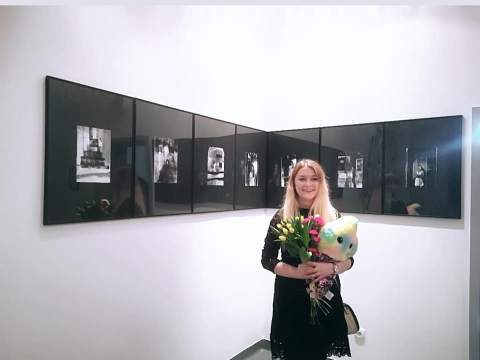 Kobieta z bukietem kwiatów i maskotką w rękach stoi na tle ściany z wiszącymi obrazami.