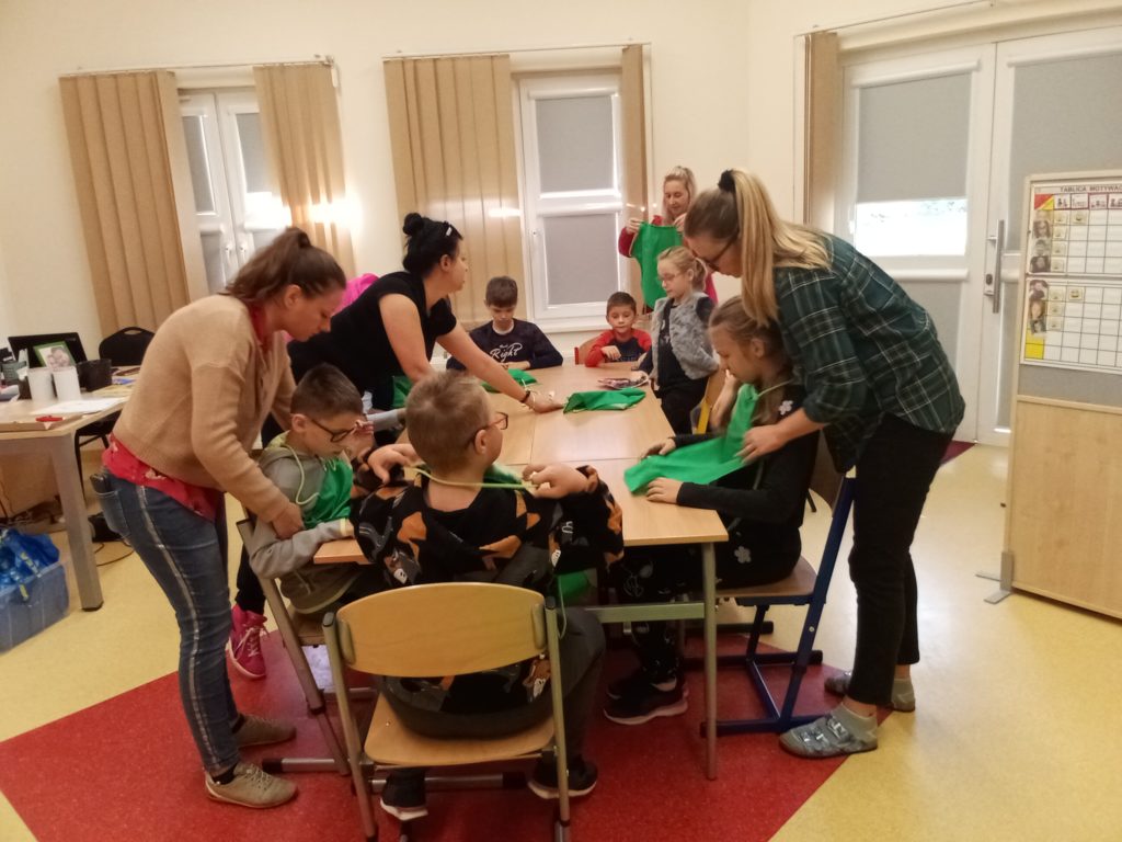 Siedmioro dzieci siedzi przy długim stole, 3 nauczycielki i instruktorka pomagają dzieciom założyć zielone fartuszki.