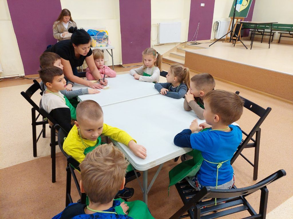 Grupa dzieci siedzi na auli przy białym stoliku na krzesełkach. Prowadząca wpisuje na samoprzylepnych karteczkach imiona dzieci. W tle opiekunka grupy, wejście na scenę.
