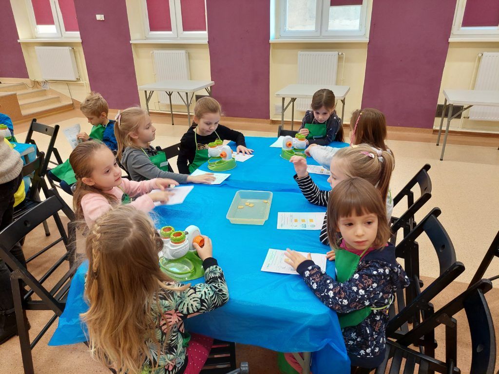 Aula, grupa ośmiu dziewczynek siedzących przy stole pokrytym niebieską folią. Na stole trzy mikroskopy, pojemnik na odpady. Przed sobą dzieci mają karty pracy na które przyklejają sól. W tle trzy małe białe stoliki.