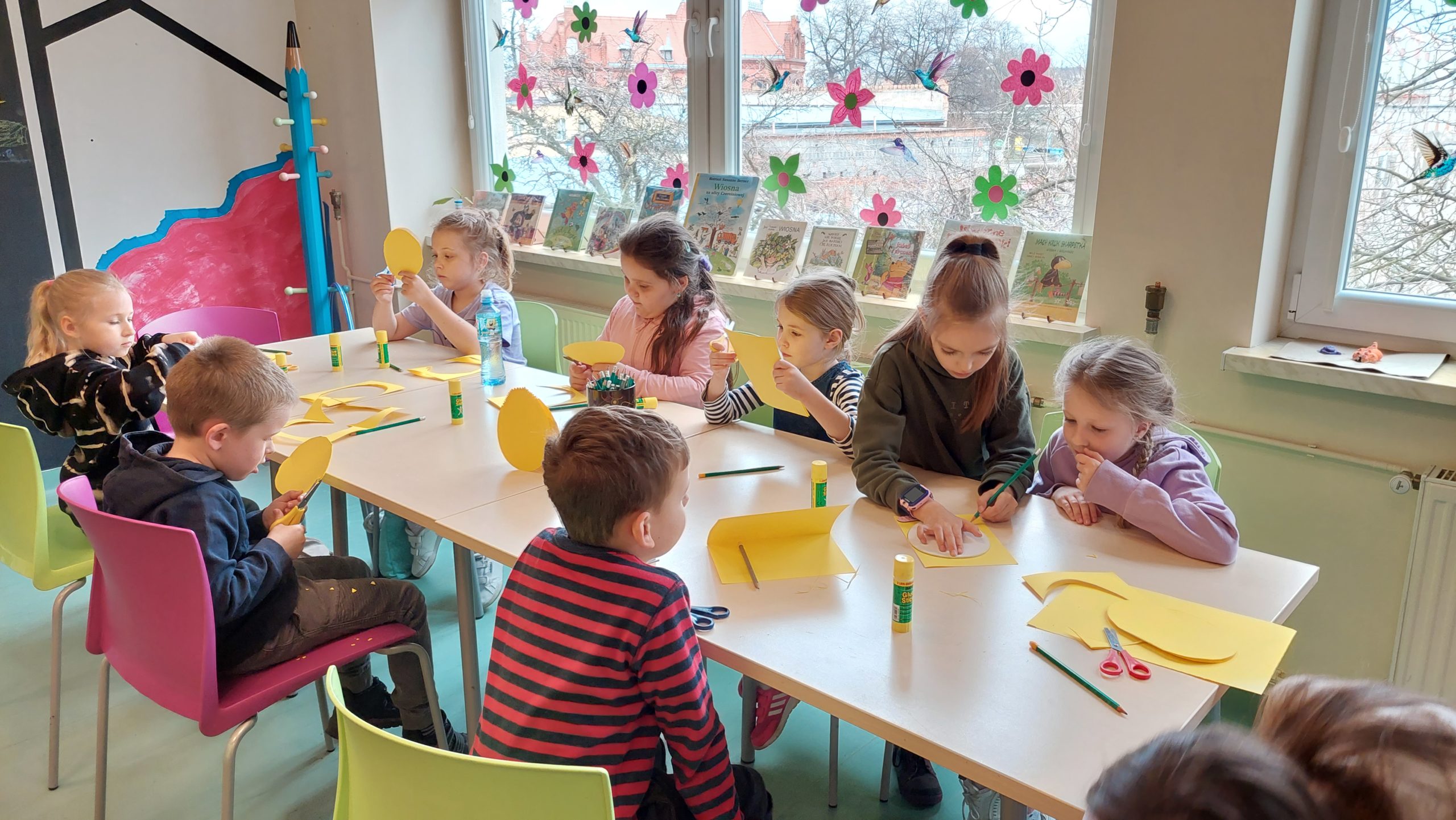 Czytelnia dla dzieci. Siedzące przy stołach dzieci odrysowują na żółtej kartce kształt jajka i wycinają.