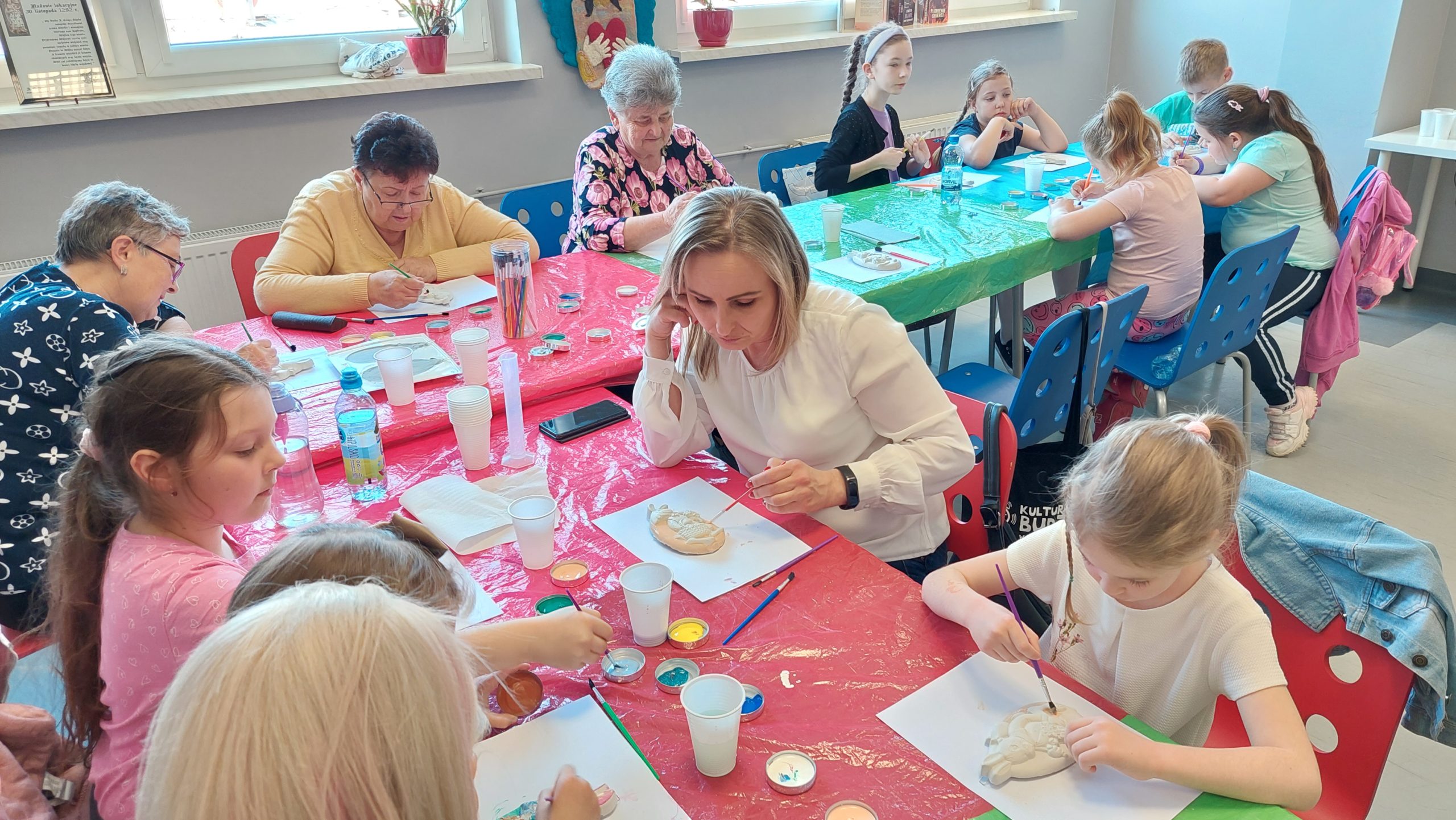 Czytelnia biblioteki. Dorośli, dzieci i seniorzy przy stołach malują farbami gipsowe zawieszki w kształcie jajka.