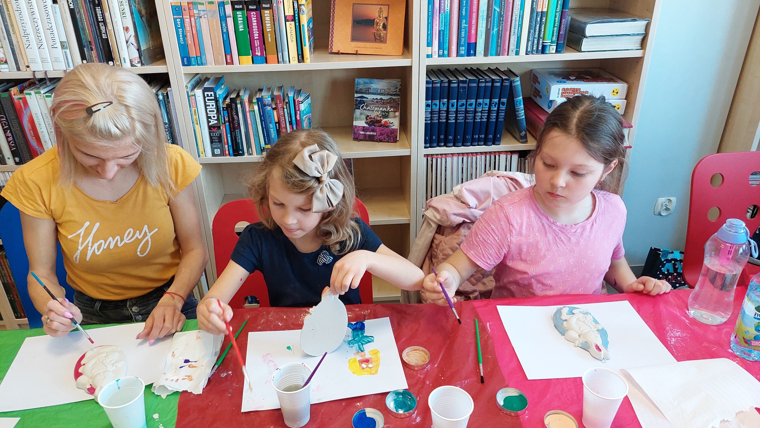 Czytelnia biblioteki. Kobieta i dwie dziewczynki malują gipsowe jajko z króliczkiem kolorowymi farbami.
