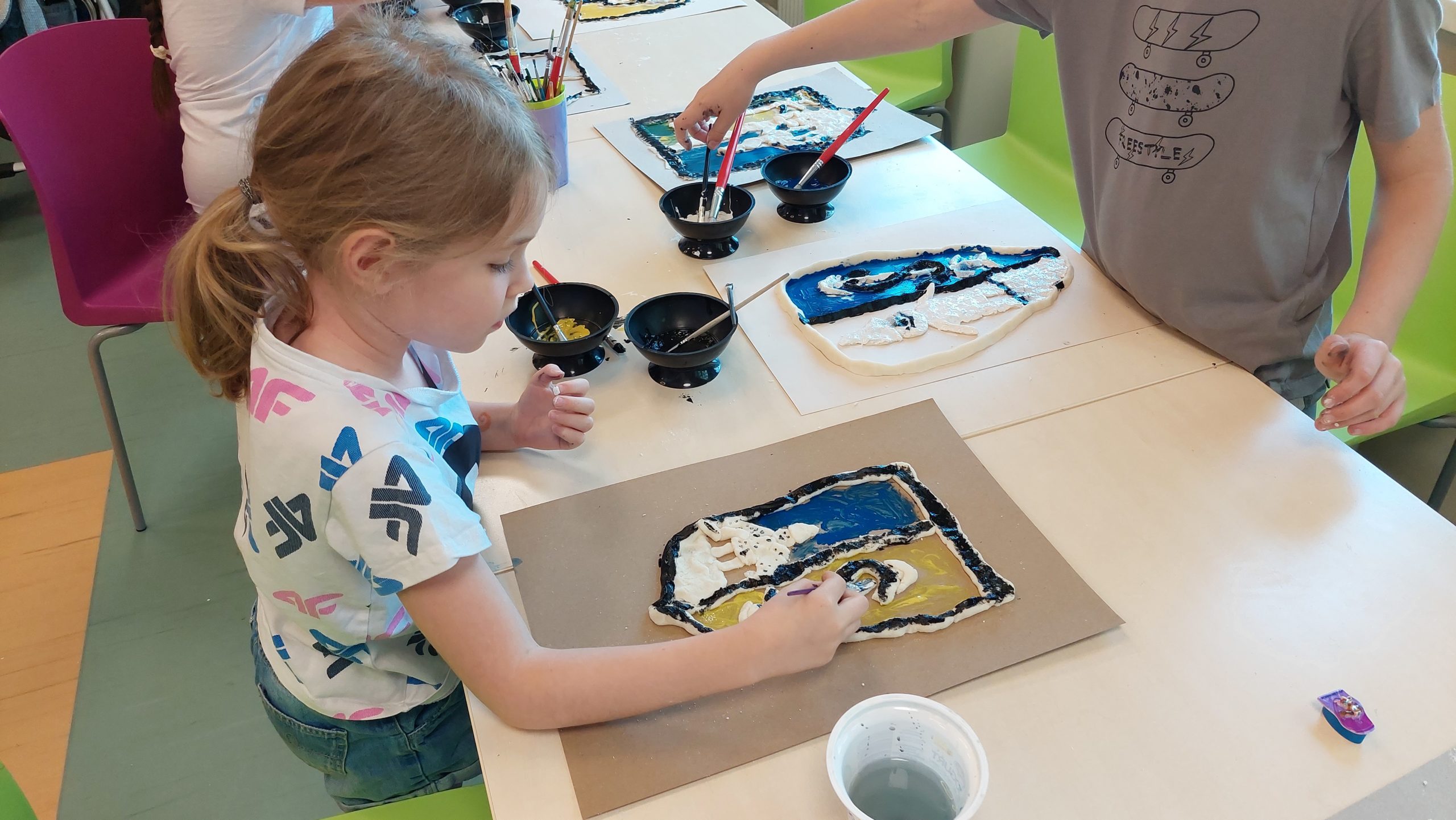Czytelnia. Dziewczynka stoi przy stole i kończy malowanie farbami swojego herbu. Na stole pojemniki z farbami, pędzle.