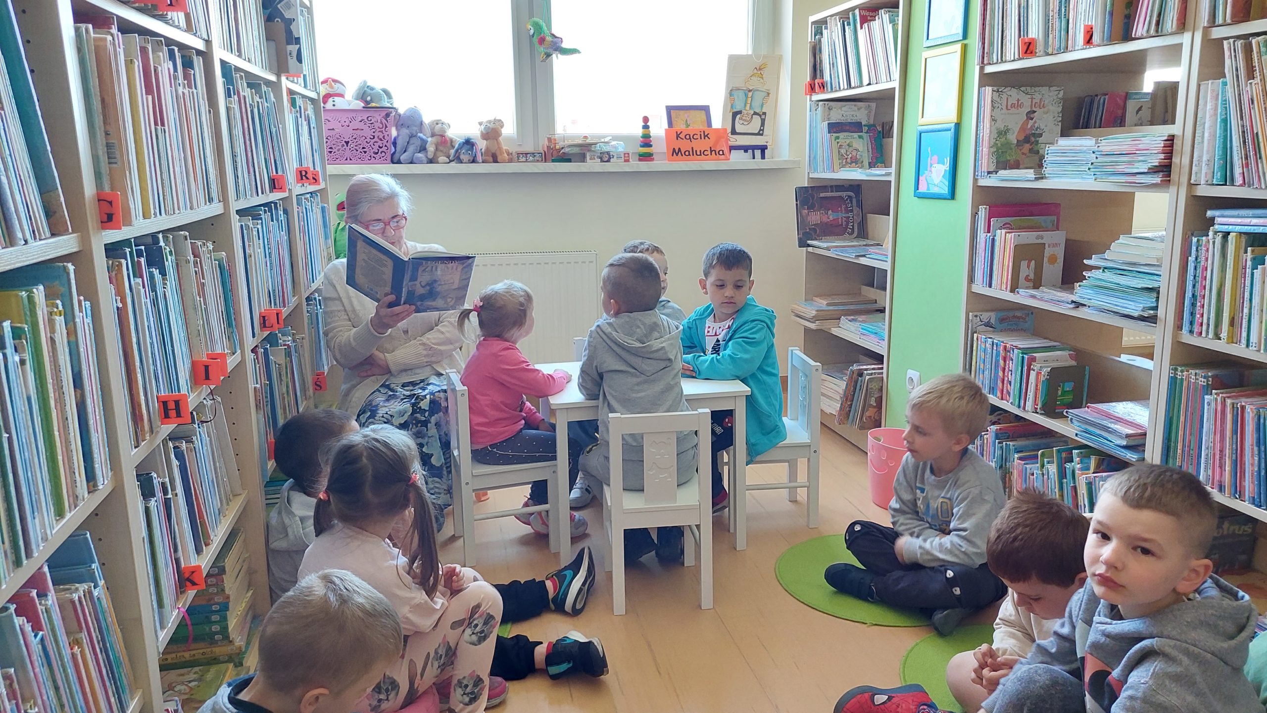 Kącik malucha. Dzieci siedzą na krzesełkach i dywanikach, obok kobieta czyta legendę z książki.