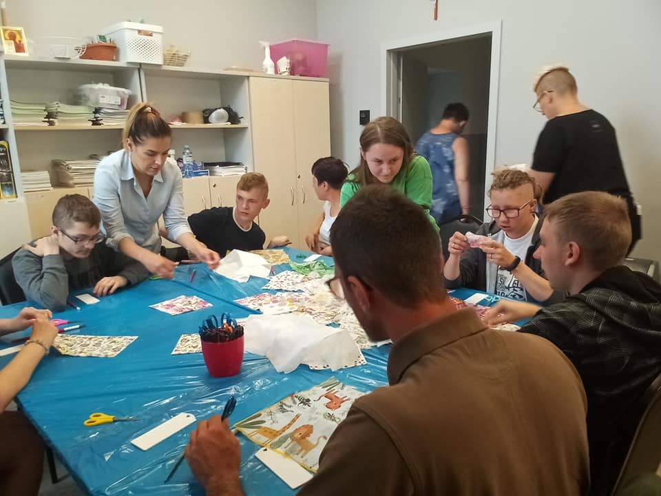 Warsztaty decoupage. Stowarzyszenie św. Celestyna w Mikoszowie -sala warsztatowa.Młodzież siedzi przy wspólnym stole , na którym rozłożone są materiały do pracy (serwetki,drewniane zakładki, klej, pędzle i nożyczki)