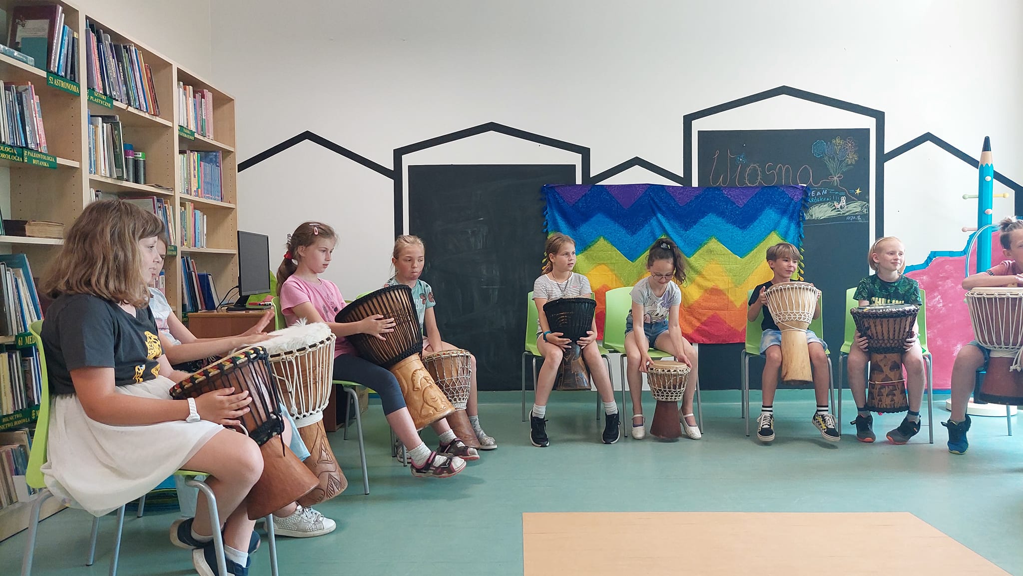 W czytelni siedzi grupa dzieci i pod okiem prowadzącego graja na afrykańskich bębnach.