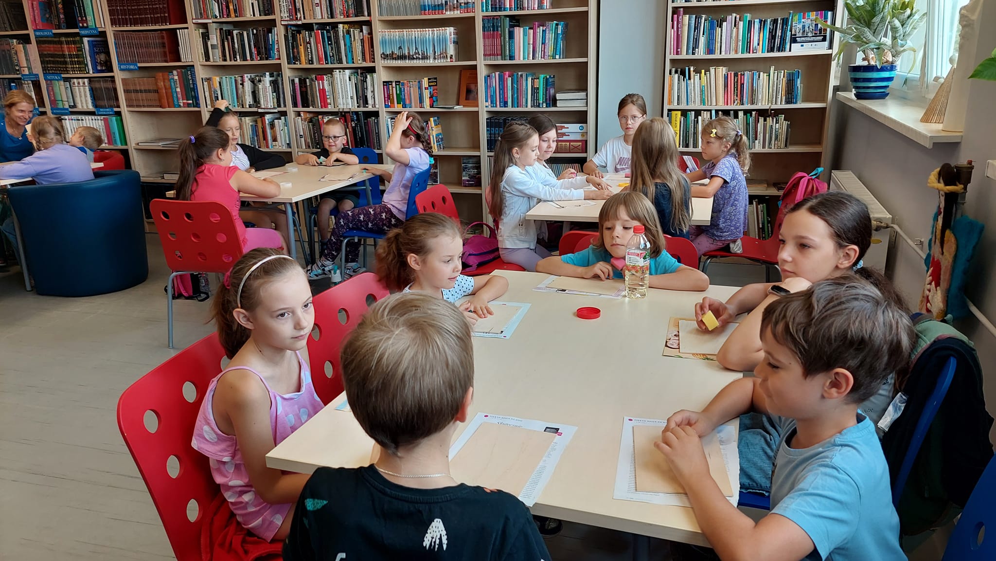 Dzieci podzielone na grupy siedzą przy stołach, na których leżą deseczki do wykonania obrazka. W ręku trzymają kawałki gąbki.