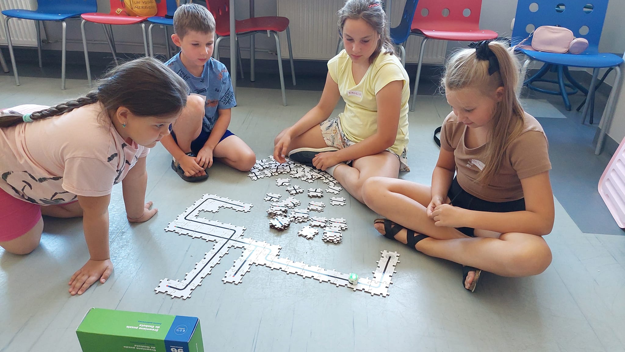 Biblioteka, czytelnia dla dorosłych. Dzieci siedzi na podłodze w grupach i rozbudowują tor w kształcie puzzli, po którym przemieszcza się mały robot.