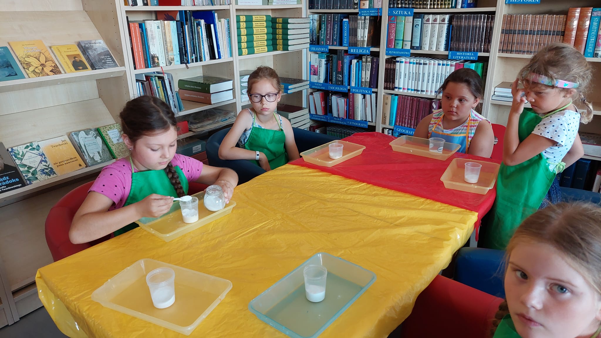 Przy stole siedzą dziewczynki w fartuszkach. Leża przed nimi pojemniczki plastikowe ,do których wsypują sól .