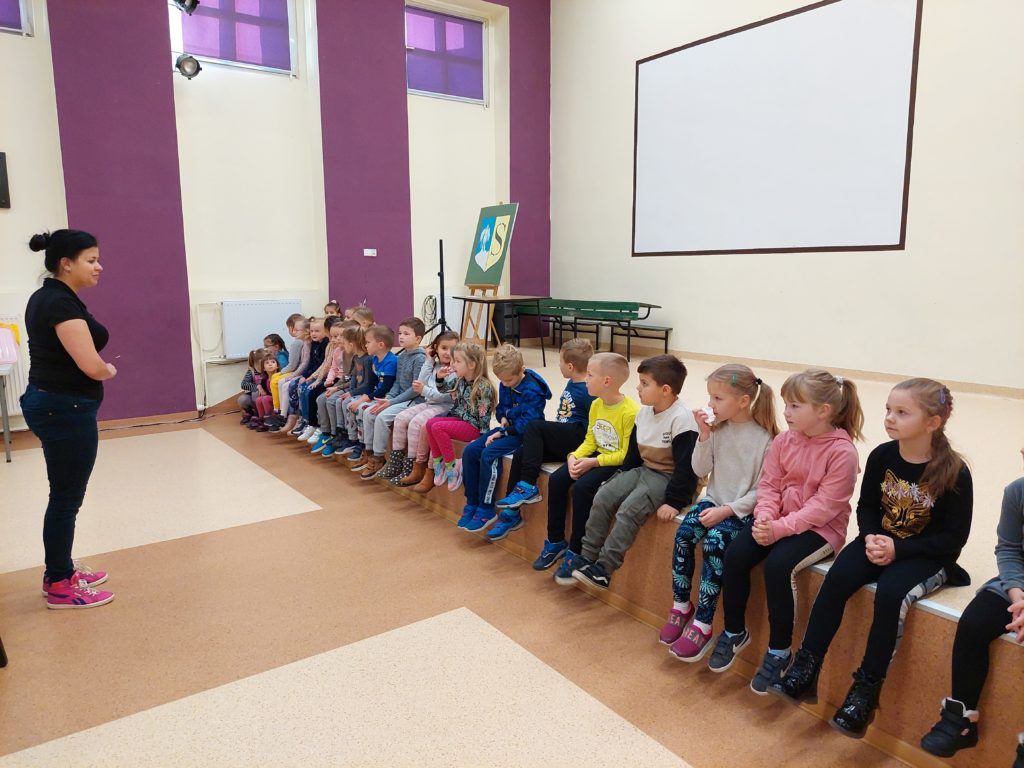 Grupa 5-6 latków siedzi na scenie auli Szkoły Podstawowej nr 3. Pani Daria stoi naprzeciw dzieci i je wita.