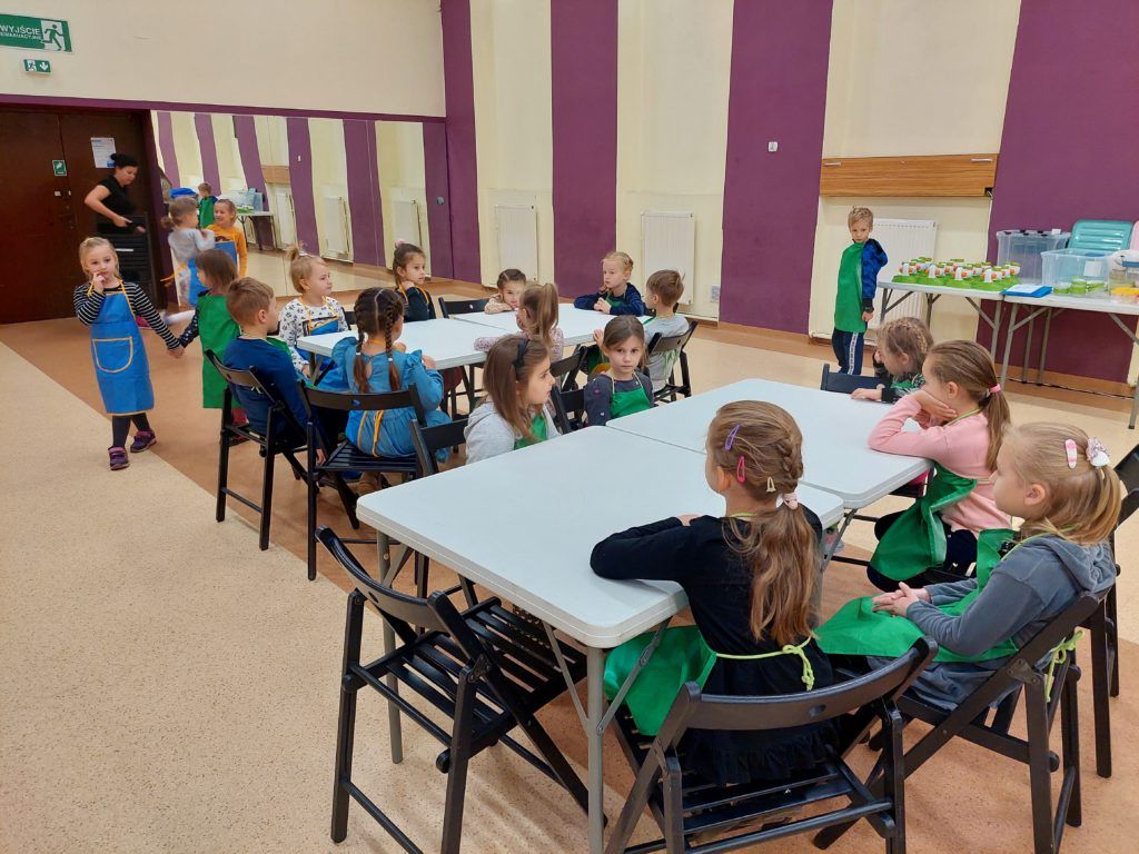Na środku sali stoją dwa duże stoły, wokół których siedzą ubrane w zielone fartuszki dzieci. W tle widać stolik z akcesoriami do zajęć.