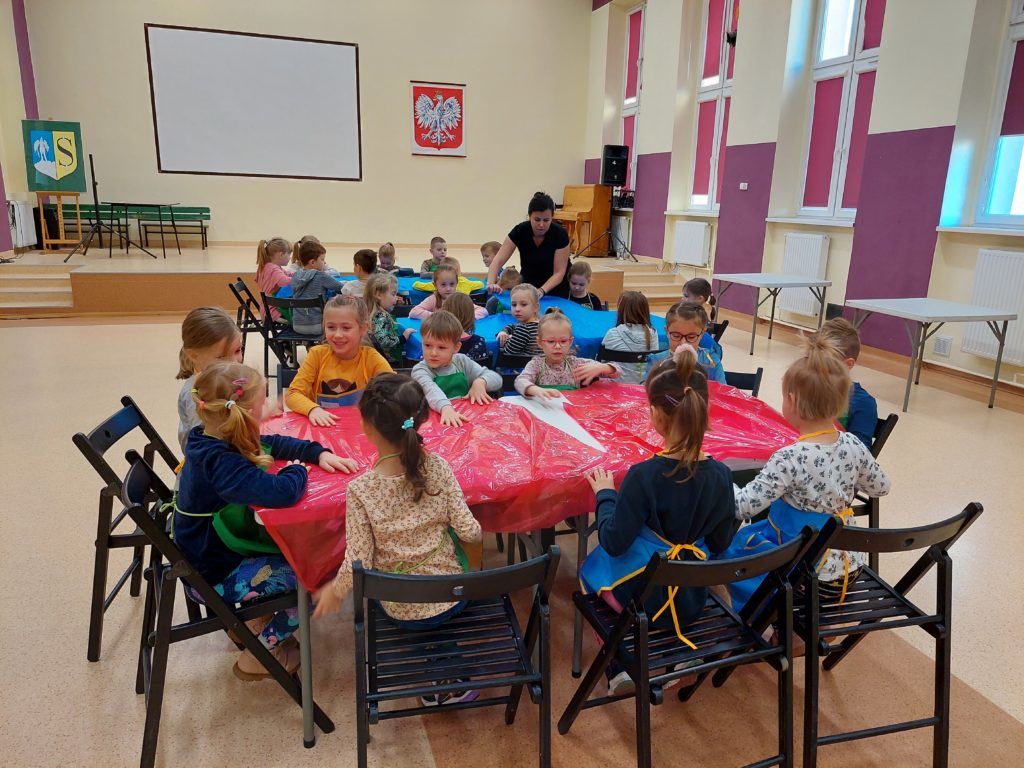 Aula, na środku stoją trzy duże białe stoły. Siedzące przy nich dzieci przykrywają je niebieską i czerwoną folią. W tle widać scenę, zawieszony na ścianie ekran, herb Strzelina oraz Godło Polski.