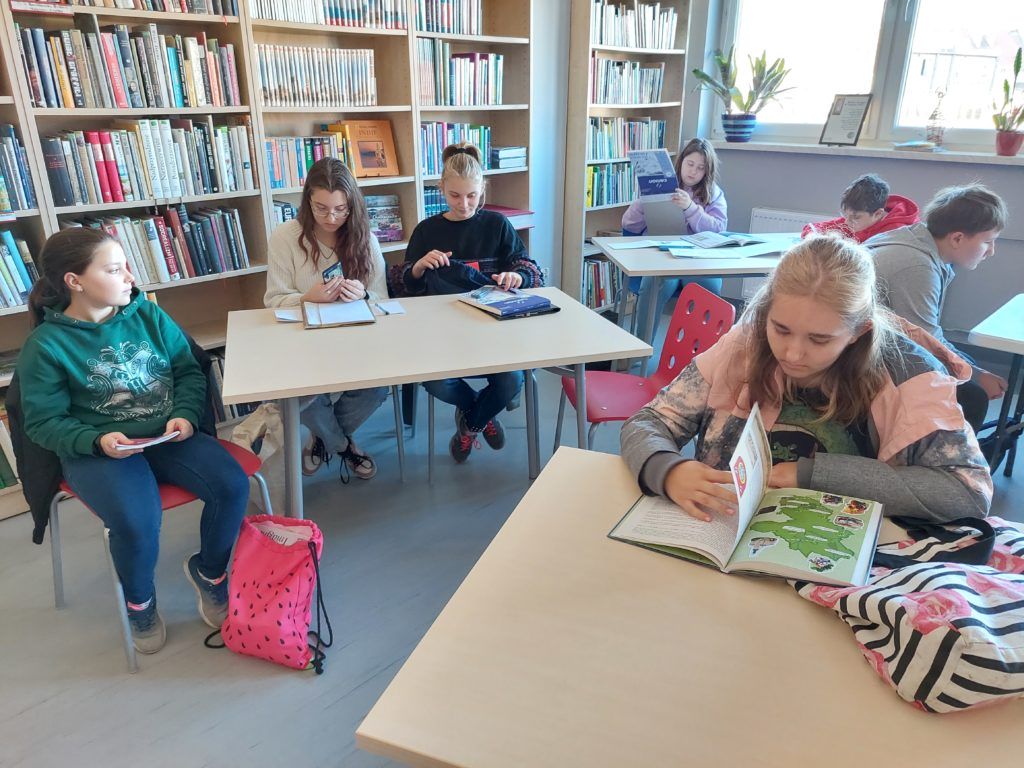 Czytelnia biblioteki, uczestnicy warsztatów siedzą przy stołach i oglądają książki z legendami o Strzelinie.
