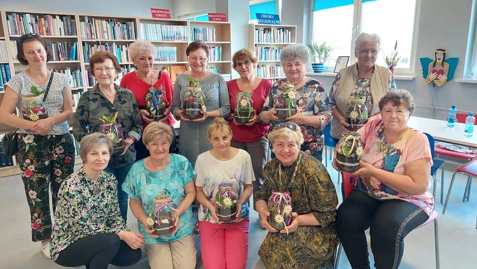 Warsztaty, czytelnia, 12 kobiet pozuje do zdjęcia i prezentuje wykonaną dekorację z żywych kwiatów "Las w słoiku"