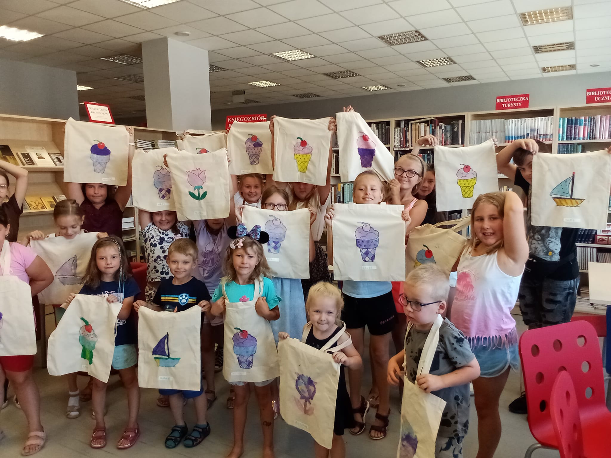 Wszystkie dzieci stoją w czytelni bibliotecznej dla dorosłych , prezentując wykonane torby.