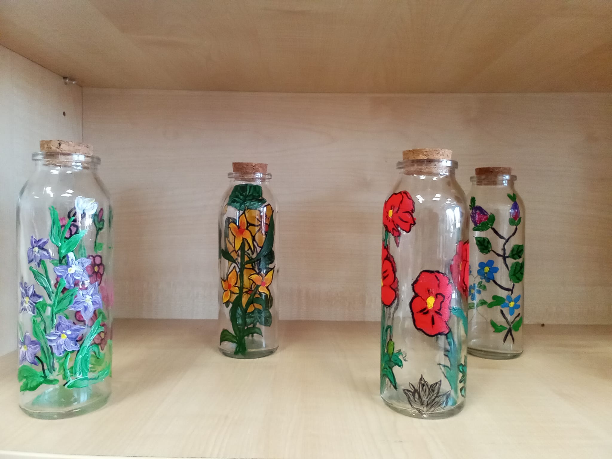Na regale stoją 4 buteleczki, malowane różnymi kolorami farb. Na wszystkich występuje motyw kwiatowy.