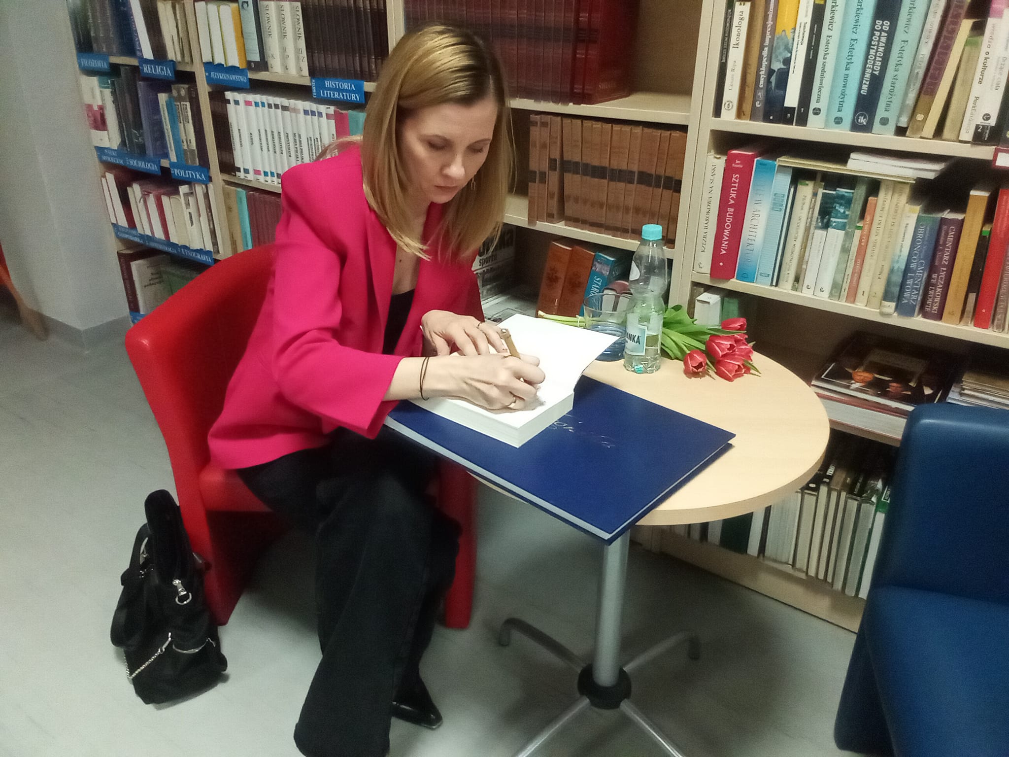 Czytelnia biblioteki, spotkanie autorskie z Joanną Opiat-Bojarską, kobieta siedzi w fotelu i wpisuje się do księgi pamiątkowej