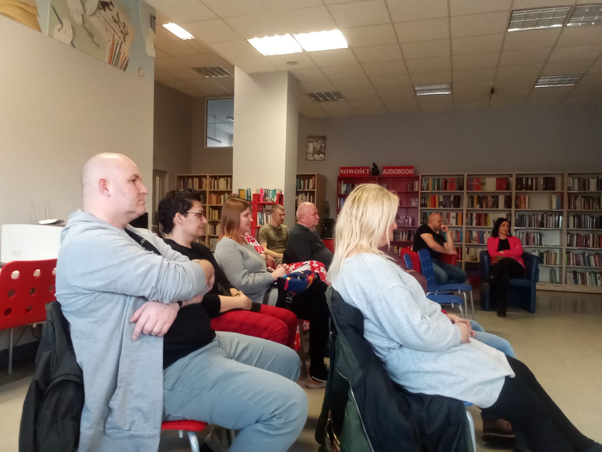 Czytelnia biblioteki, spotkanie autorskie z Joanną Opiat-Bojarską,grupa osób siedzi na krzesłach i słucha wypowiedzi autorki