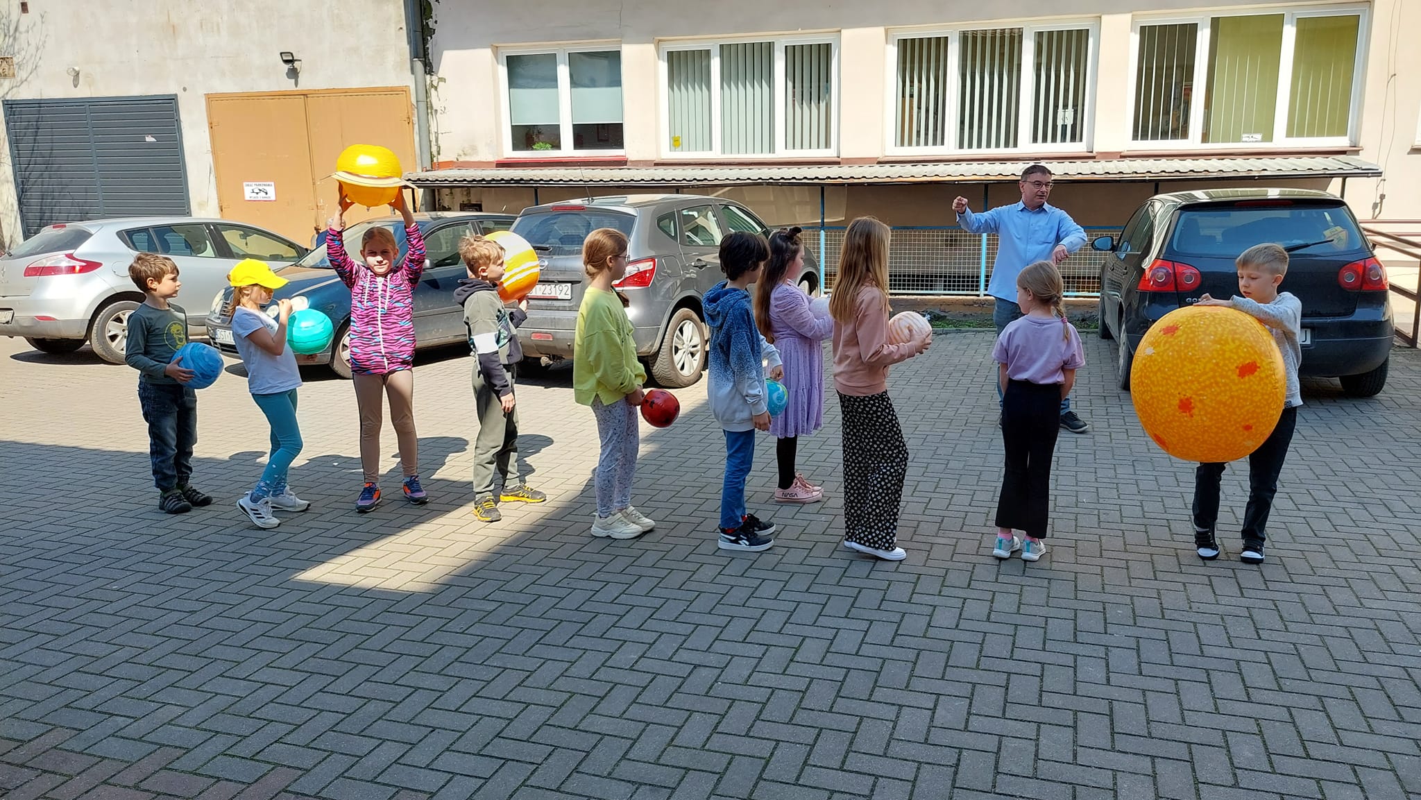 Parking przed biblioteką. Grupa dzieci z dmuchanymi planetami odtwarzają Układ Słoneczny pod okiem prowadzącego