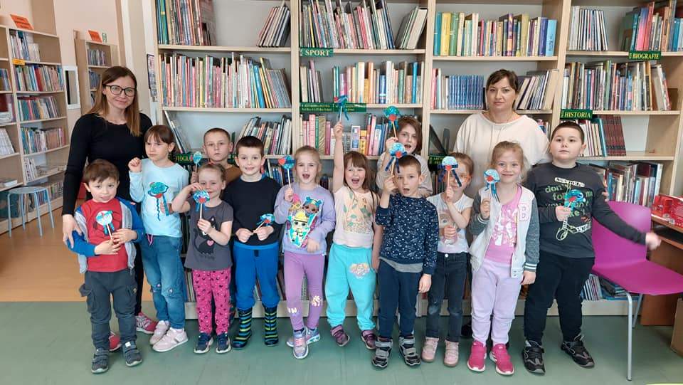 Czytelnia dla dzieci. Grupa przedszkolaków i dwie panie pokazują wykonane mini kule ziemskie na patyczku