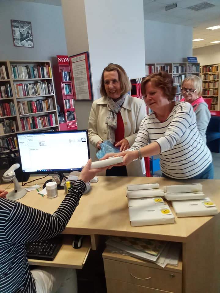 Trzy kobiety stoją przed ladą biblioteczną jedna z nich podaje bibliotekarce owiniętą w papier książkę