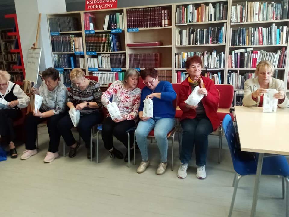 Czytelnia biblioteki, siedem pań siedzi na krzesłach, każda trzyma w rękach torebeczkę z prezentami.