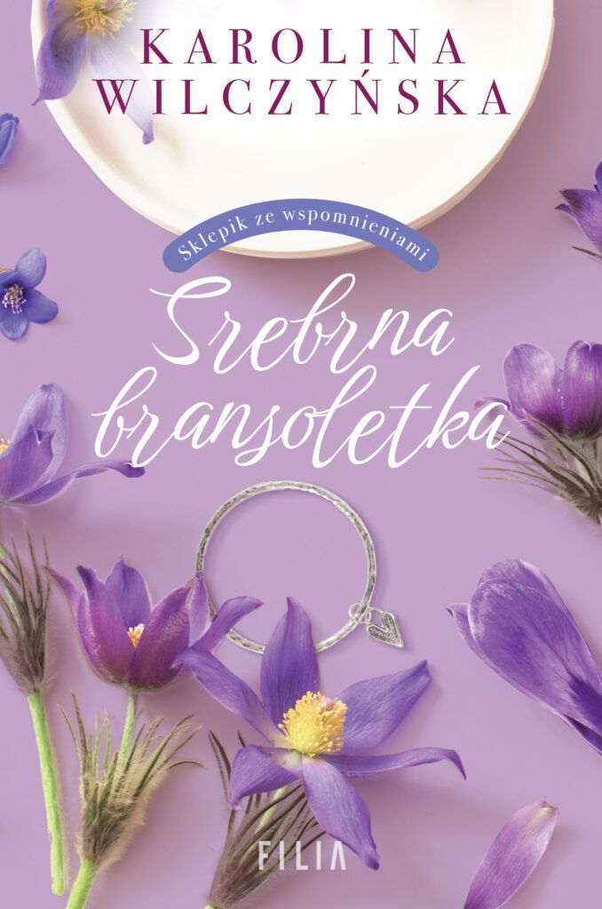 Na liliowym tle fragment białego talerza, fioletowe kwiaty oraz srebrna bransoletka z serduszkiem